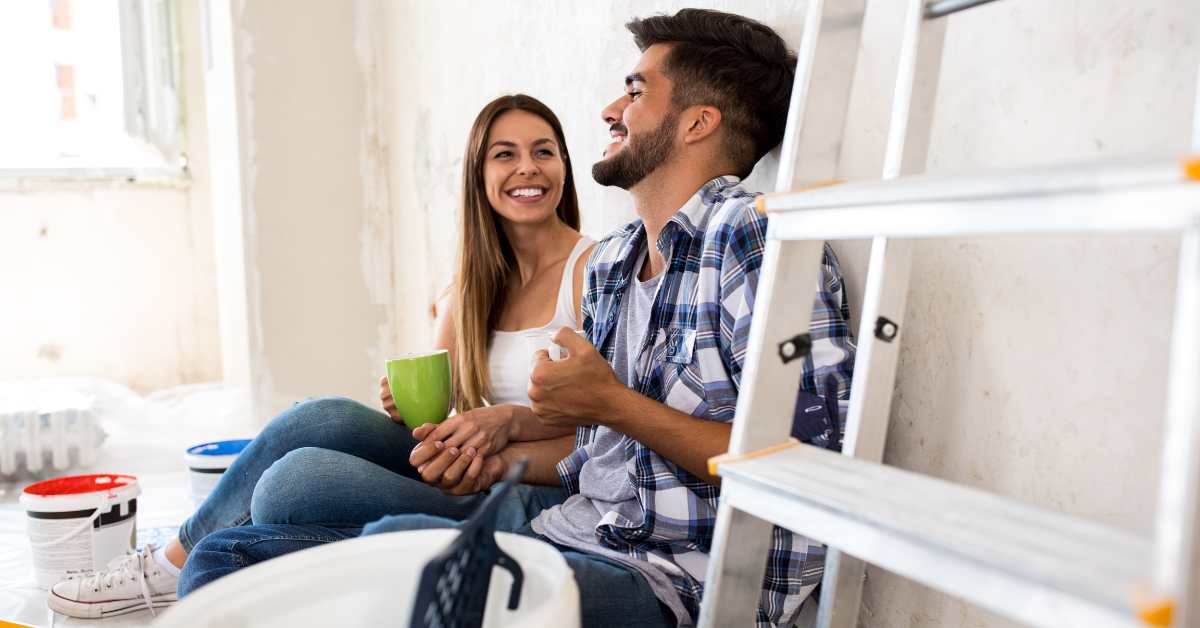 A happy young couple sitting on the floor of their Scottsdale home with paint buckets and a ladder next to them.